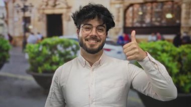 Gözlüklü ve sakallı İspanyol genç adam kalabalık bir sokakta dostça ve olumlu bir hava sergiliyor..