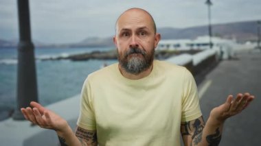 Deniz kenarındaki gezinti alanında sakallı bir adam sıradan sarı gömlekli, kolları dövmeli, deniz zeminine yaslanmış, açık havada yaz havası yayıyor ve düşünceli bir ifade takınıyor..