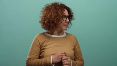 Woman with glasses standing against a green wall with a pensive expression in a studio setting wearing a brown sweater and looking away from the camera