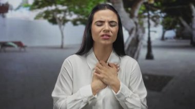 Parkta göğsü ağrıyan bir kadın, endişeli bir ifadeyle, beyaz gömlek giyiyor, elleri göğsüne yapışıyor, arka planda ağaçlar var, rahatsızlık ve sıkıntı taşıyor..