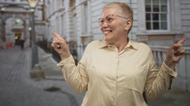 Gözlüklü ve bej tişörtlü bir kadın, gösterişli bina cephesinin yanında, caddenin her iki tarafına işaret parmaklarıyla işaret ediyor..