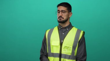 Gözlüklü ve yansıtıcı yelekli genç adam sağlam yeşil bir zemine karşı duruyor, düşünceli bir ifade sergiliyor, inşaat ve güvenlik temaları vurguluyor.