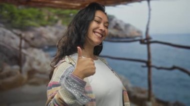 Kadın parmaklarını kaldırır ve kayalık sahilde tahta korkulukların yanında kameraya işaret eder; iyimserlik neşe teşviki.