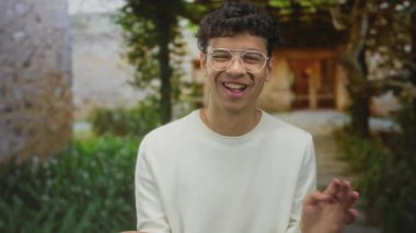 Man wearing white sweater and glasses laughs and waves hand at stone building entrance with garden pathway; joy.