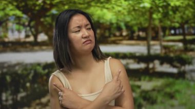 Parkta kollarını kavuşturan genç Çinli bir kadın hoşnutsuz bir ifadeyle yeşil ağaçların altında dikilirken belirsizliği yansıtıyor..