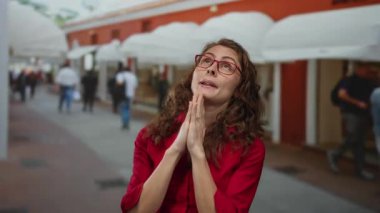 Gözlüklü ve kırmızı gömlekli bir kadın dua ederken el ele tutuşuyor. Meşgul şehir caddesinde insanlar yürüyor ve arka planda kırmızı bina var..