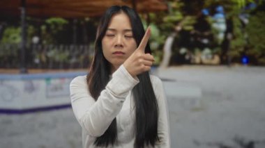 Beyaz tişörtlü Çinli kadın yeşil park ortamında saygısızca jest yapıyor..