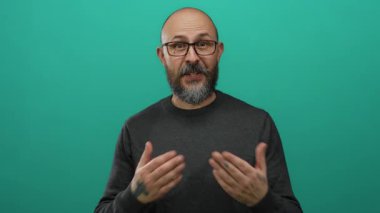 Kel ve sakallı bir adam, yeşil arka plana karşı elleri ve kalbinin üstünde şükranlarını sunuyor, gözlük takıyor ve ellerinde dövmeler gösteriyor..