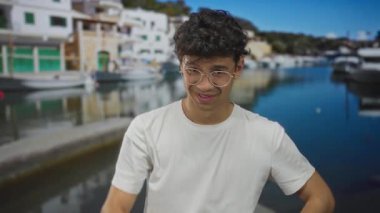 Beyaz tişörtlü ve gözlüklü bir adam limanda alnına jest yapıyor. Görünür, güler yüzlü, şakacı..