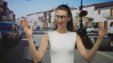 İki elini kaldıran kadın, avuçlarını güneş ışığıyla aydınlatan bir caddede, gözlüklü ve kolsuz beyaz bir bluz giyerken ağız açık ve gözler geniş; oyuncu sürpriz.