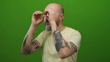 Dövmeli ve sakallı kel bir adam canlı yeşil bir arka planda hayali dürbünlere bakarak neşeli ve izole bir atmosfer yaratıyor..