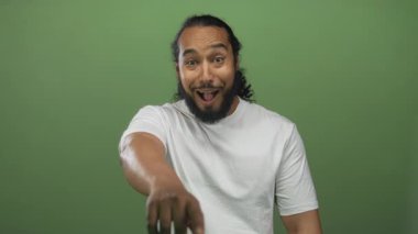 Elleri yanaklarda, ağzı açık, gözleri açık, şaşırmış bir adam stüdyoda yeşil arka planda oturuyor; sürpriz tepki.