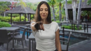 Genç kadın, çardak ve sandalyeli havuz kenarındaki yemek alanındaki kamerayı işaret ediyor..