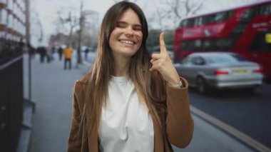 Arka planda kırmızı çift katlı otobüsü olan, uzun saçlı, gülümseyen, kahverengi ceketli ve beyaz gömlekli genç bir kadın..