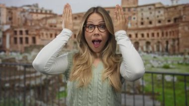 Yuvarlak gözlüklü sarışın kadın, gülümseyen yüzünü Roma harabelerinin ortasında elleriyle çerçeveliyor..