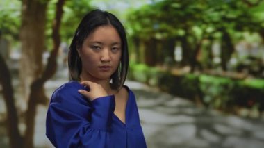 Ağaçlarla çevrili, açık havada, yemyeşil bir parkta, mavi tişörtlü bir kadın, çeşitli duygular ifade ediyor, sakin bir zemin ve yumuşak ışıklandırmayla doğanın sükunetini yakalıyor..