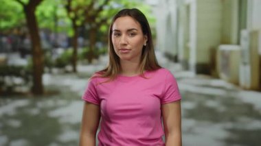 Pembe tişörtlü bir kadın güneşli bir günde ağaçlarla kaplı bir sokakta dikiliyor..