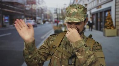 Sırt çantası ve künyeli kamuflaj üniforması giyen adam kalabalık bir caddede burnunu çimdikliyor..