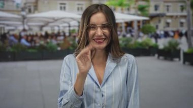 Gözlüklü bir kadın açık hava restoranının yanındaki bir kafede otururken gülümsüyor ve yumruğunu kaldırıyor..