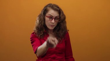 Kıvırcık saçlı ve kırmızı gözlüklü bir kadın. Öfkeyle turuncu bir arka plana işaret ediyor. İddialı bir hava yaratıyor.