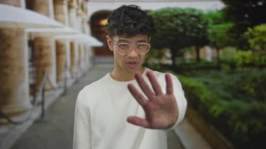 Beyaz kazaklı adam bir binadaki taş sütunların arasında avuç içi kamerayı ileri doğru tutar; reddedilme.