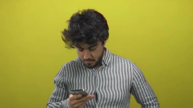 Sakallı genç adam, parlak sarı arka plana karşı akıllı telefon mesajı dinliyor. Modern iletişim ve rahat bir yaşam tarzı öneriyor..