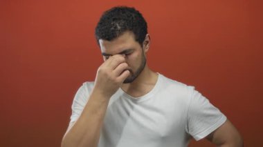 Turuncu stüdyoda parmak ve işaret parmağıyla burnunu çimdikleyen bir adam beyaz tişört giyer ve yüz yorgunluğu gösterir..