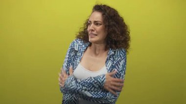 Sarı duvarlı bir stüdyoda çapraz kollar ve gergin omuzlarla kendini kucaklayan genç bir kadın..