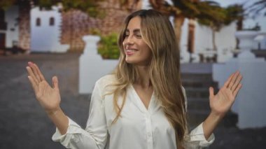 Beyaz bluz giyen genç sarışın kadın güneşli bir avluda el büyüklüğünde jest yapıyor..