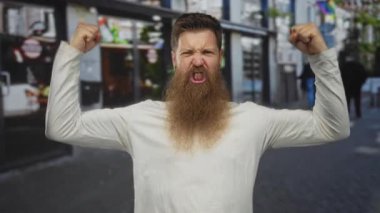 Sakallı genç adam, dükkan önleri yakınındaki kalabalık şehir sokağında kaslı yumruklarıyla kaslarını esnetiyor, dişlerini sıkıyor;.