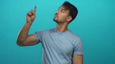 İzole edilmiş mavi bir arka planda el kol hareketi yapan genç bir İspanyol, kendine güvenen bir ifadeyle yukarıya doğru işaret ediyor ve açık mavi bir gömlek giyiyor..