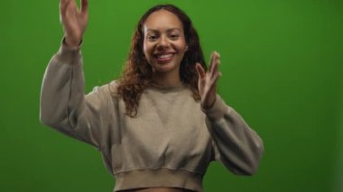 Kırpılmış bir kazak giyerken yüzünü elleriyle çerçeveleyen ve stüdyoda yeşil bir zemine gülümseyen bir kadın; keyif güveni..