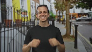 Siyah tişörtlü adam gülerek caddede dişlerini gösteriyor demir korkuluklar ve ağaçlar; mutluluk güveni.