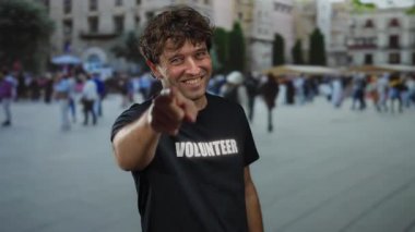 Gönüllü tişört giyen İspanyol adam işlek caddede gülümseyip el kol hareketleri yapıyor. Şehir ortamında samimi ve iç açıcı bir anı yakalıyor..