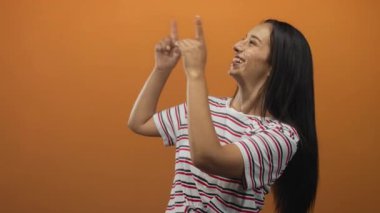 Turuncu bir stüdyoda çizgili tişört giyerek parmaklarını yukarı gösteren bir kadın..