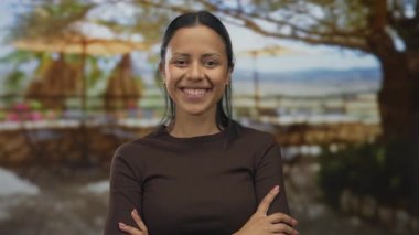 Güneşli bir günde ağaçların altında kolları çaprazlanmış, güvenli bir şekilde gülümseyen İspanyol genç bir kadın. Renkli çiçeklerin bulanık arka planda zarifçe açtığı yerde..