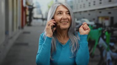 Gri saçlı, mavi kazaklı yaşlı bir kadın, bisiklet ve şehir manzarasıyla çevrili canlı bir sokak ortamında dikilirken akıllı bir telefonla neşe içinde konuşuyor..