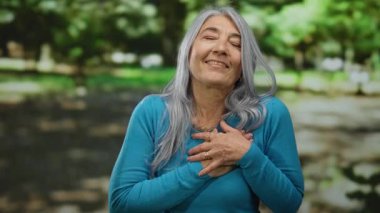 Son sınıf öğrencisi, kır saçlı, elleri göğsünde, doğa ve yeşilliklerle çevrili güneşli bir parkta minnettarlığını ifade eden, dışarıdaki huzur ve mutluluğu somutlaştıran bir kadın..