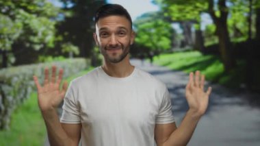 Parlak güneş ışığı altında yemyeşil bir parkta neşeli hareketler yapan genç adam, arka planda ağaçlar ve patikalarla tasasız ve neşeli bir hava yayıyor..