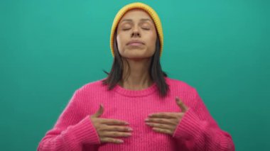 Pembe kazak ve turuncu şapka giyen, gözleri kapalı, sakin nefes alan bir kadın izole bir ortamda sükunet ve rahatlık sergiliyor..