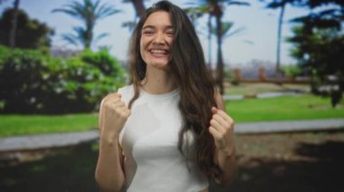 Kadın, yeşil parktaki palmiye ağaçları arasında güneş ışığı altında yumruklarını sıkar ve gülümser; sevinç zaferi kutlamaları.