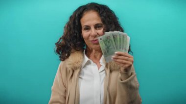 Orta yaşlı bir kadın mavi bir duvara karşı eğlenceli bir jest yaparak, neşeli ve kendinden emin bir tavır sergiliyor..