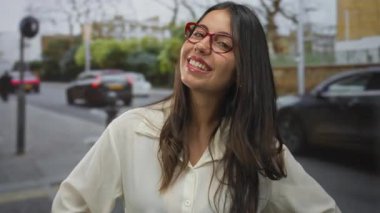 Kalabalık bir caddede kırmızı gözlüklü, beyaz gömlekli, araçlarla çevrili, gülümseyen bir kadın, Londra 'nın hareketli bölgesinde canlı bir şehir havası yakalıyor..