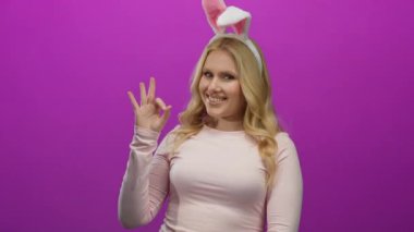 Tavşan kulağı takan ve parlak pembe arka plana karşı iyi bir hareket yapan, neşeli davranışlar sergileyen, sarışın ve genç görünümlü neşeli ifadeler sergileyen bir kadın..