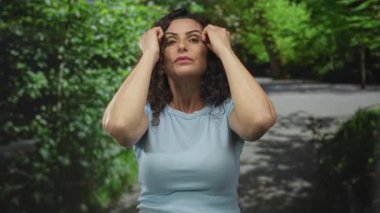 Açık mavi gömlekli genç kadın kıvırcık koyu saçlı, iki eli yeşil parkta alnına kadar kalkık; stres.