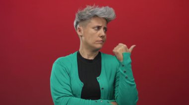 Kırmızı duvarla aydınlatılan stüdyoda gümüş rengi saç kesimi, yeşil hırka ve siyah üst başlık giyen bir kadın..