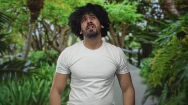 Man with hands raised and bare arms looking up on a forest path, white tshirt and visible chest; quiet hopeful reflection.