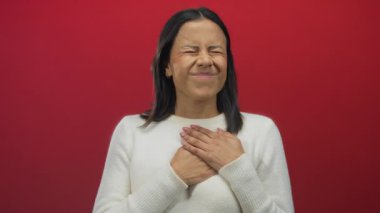 Beyaz kazaklı İspanyol kadın izole edilmiş kırmızı arka plan duvarına karşı acı içinde kalbini tutarak rahatsızlık ve duygusunu dile getiriyor..