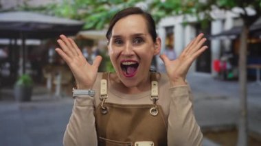 Önlüklü İspanyol kadın canlı bir sokakta kafeler ve ağaçlarla neşeyle tepki veriyor. Canlı bir şehir ortamında şaşkınlık ve keyif ifade ediyor..