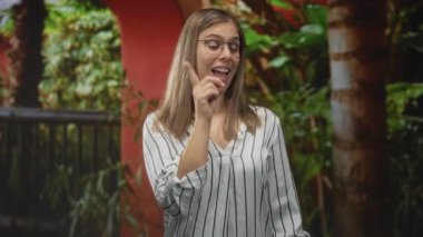 Sokakta, tropikal bitkilerin yanında, gözlük takan, çizgili bluz giyen, sarışın, eğlenceli bir kadın..